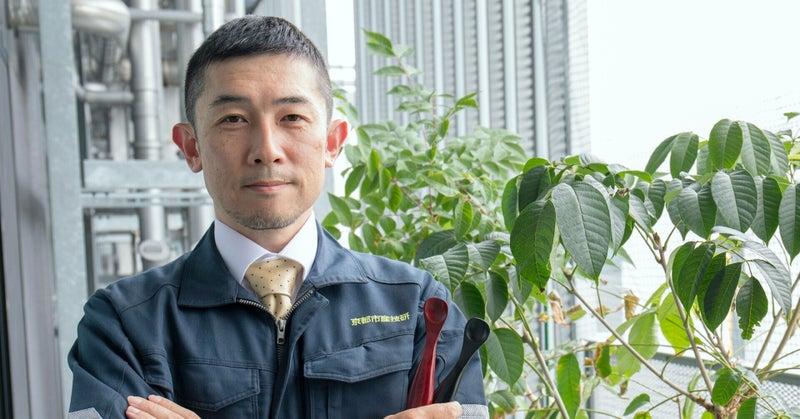 京都市産業技術研究所の竹浪祐介主席研究員　作業着を着た竹浪さんが、ご自身が開発された2本の介護スプーンを持って正面を見据えている